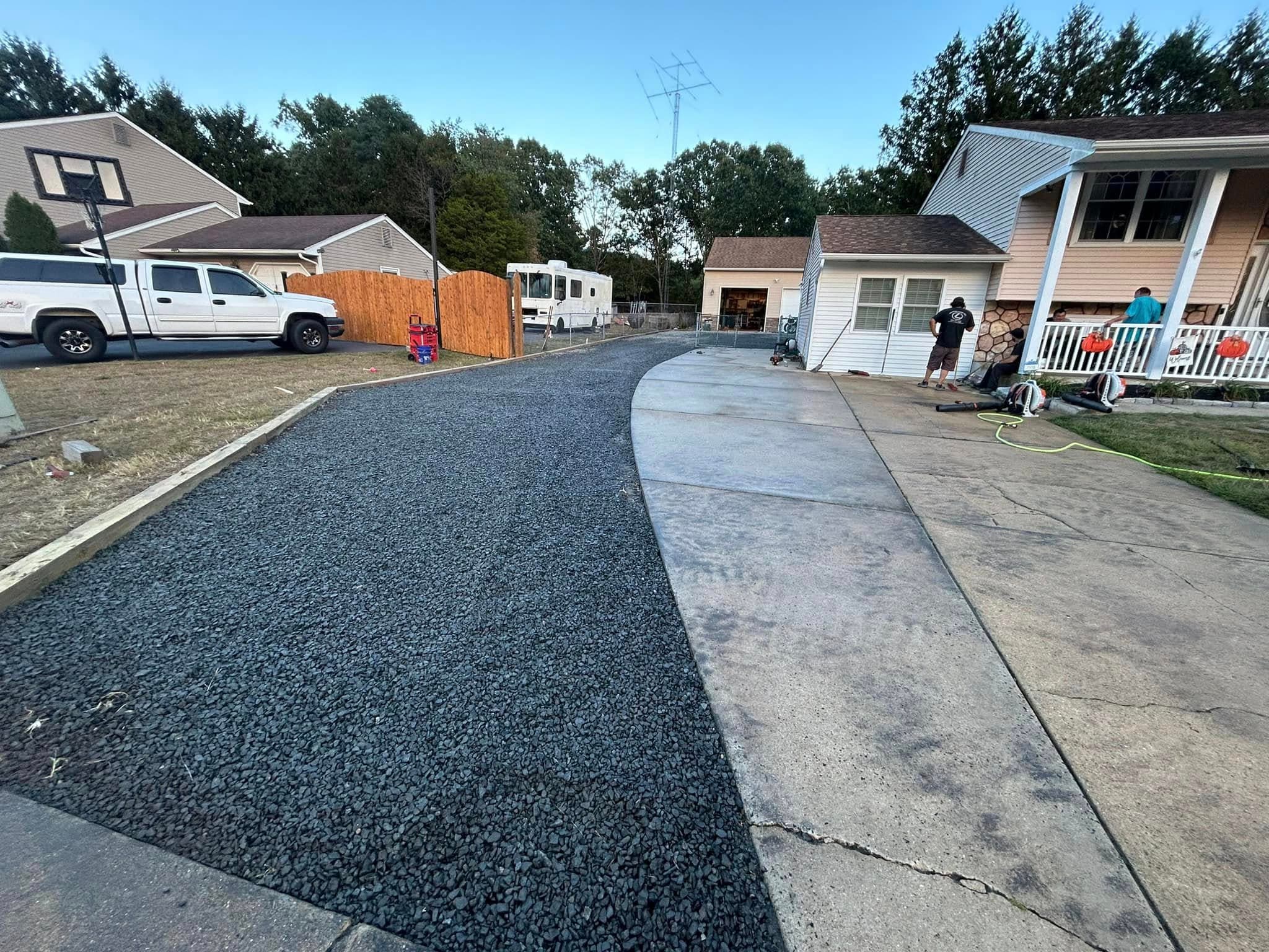 Newly installed gravel driveway and concrete area in residential neighborhood with vehicles nearby.