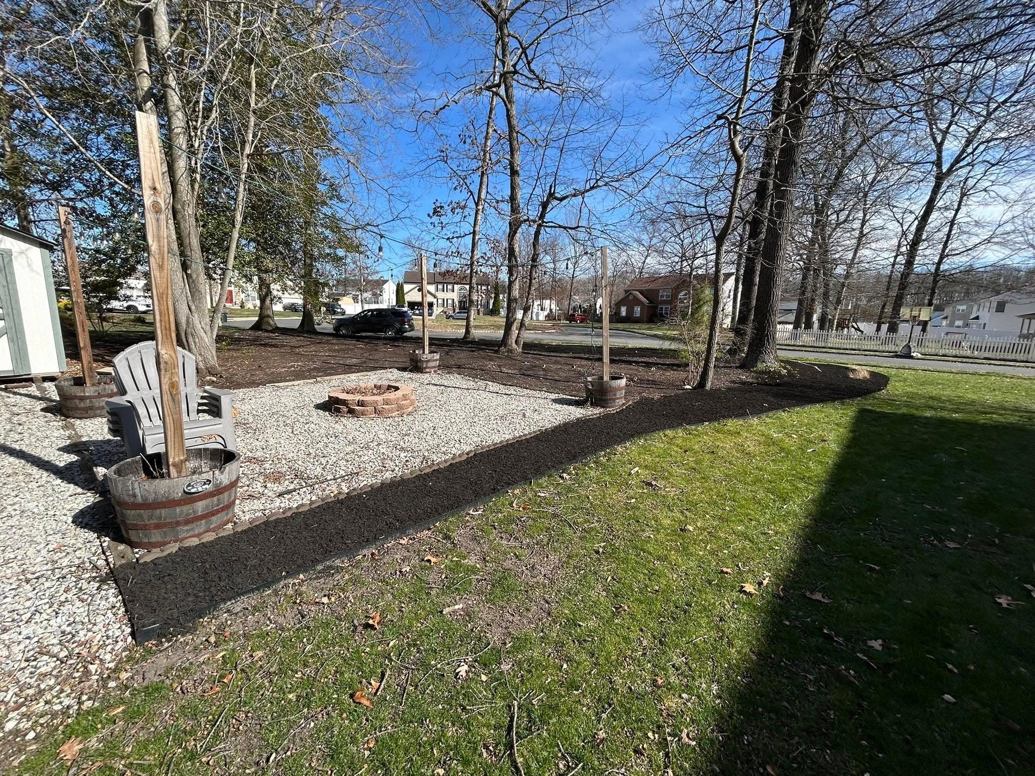 Outdoor backyard with gravel fire pit area, sitting chairs, and lush green grass under trees.